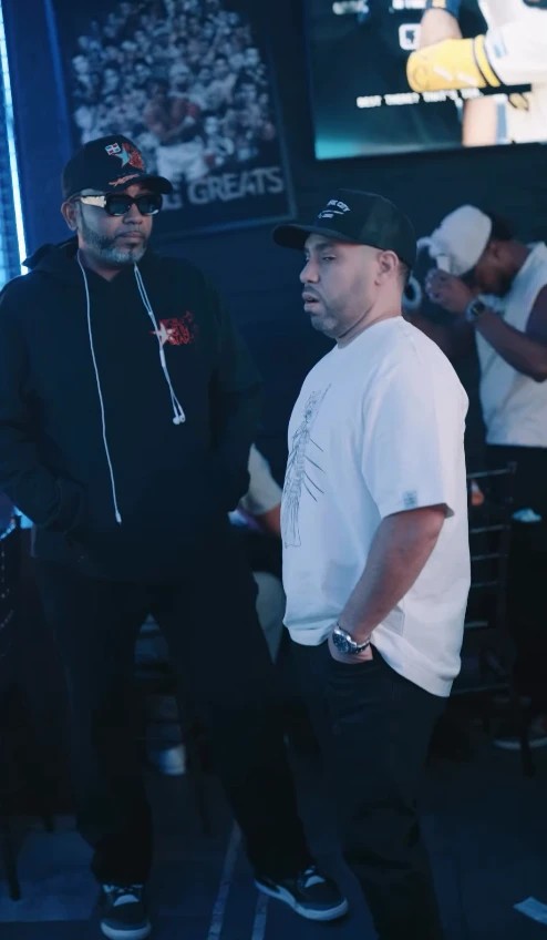 A frame from LMP's reel: two men in caps, one in a black hoodie and sunglasses, one in a white T-shirt, standing by the bar's screens