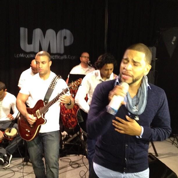 Henry Santos singing into a microphone with his band — guitar and percussion — in front of the LMP backdrop
