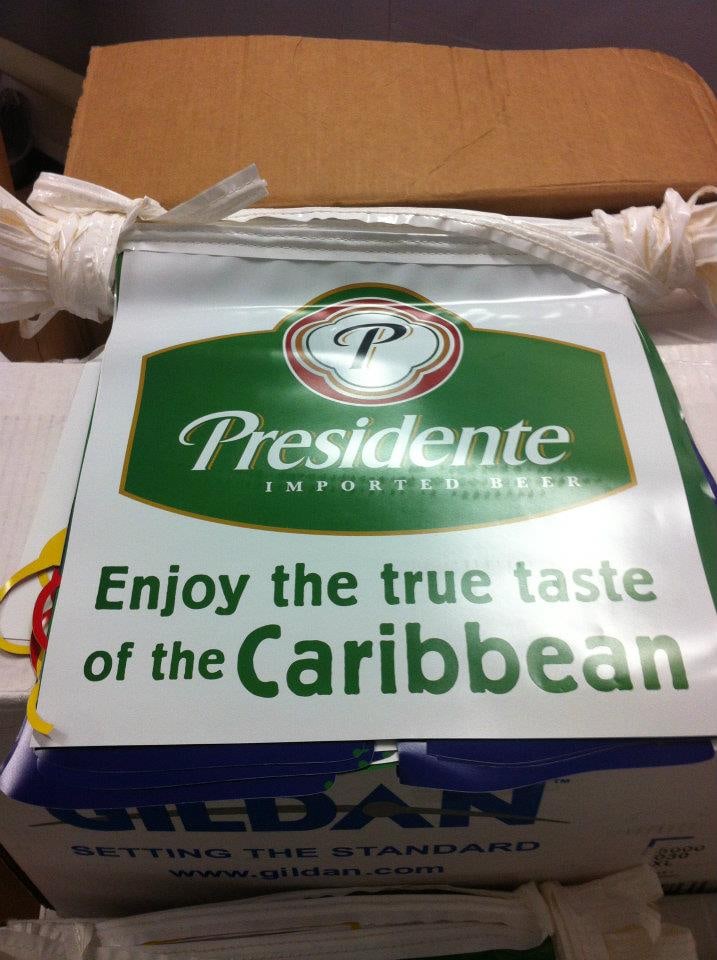 A string of Presidente “Enjoy the true taste of the Caribbean” pennant flags on a box of Gildan shirts