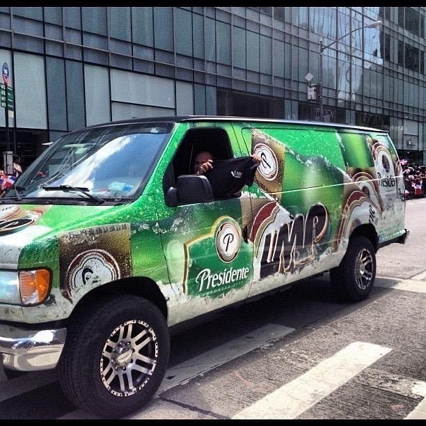 A green van wrapped in Presidente and LMP branding on a New York avenue, crowds with Dominican flags behind it