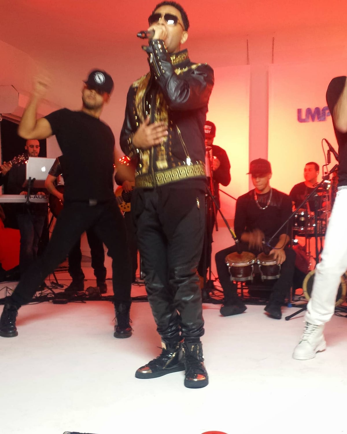 Toby Love singing in a black-and-gold studded jacket, dancers and his band around him, the LMP logo on the wall