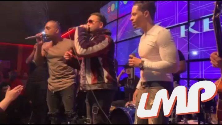 Three singers with microphones on stage, a drummer and a purple video wall behind them, the LMP logo in the corner